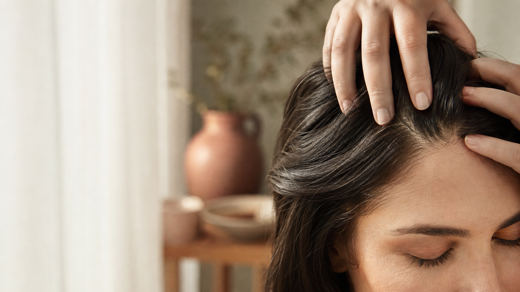 Close no alto da cabeça de uma mulher com dedos separando o cabelo escuro para expor o couro cabeludo, com jarra de cerâmica desfocada ao fundo